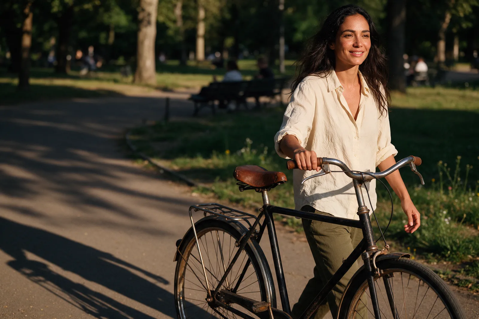 Mulher de cerca de 45 anos, de camisa clara, empurra uma bicicleta num parque e sorri, numa tarde de sol